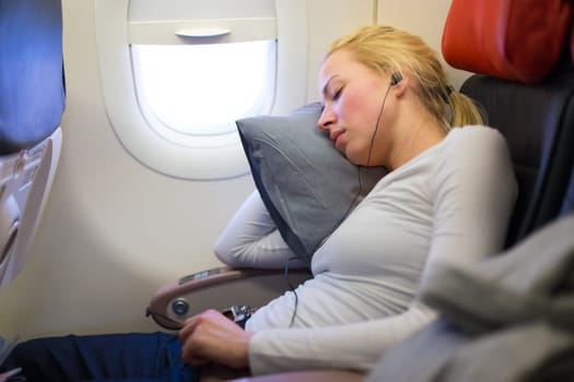 Tired blonde casual caucasian lady listens to music while napping on uncomfortable seat while traveling by airplane. Commercial transportation by planes.