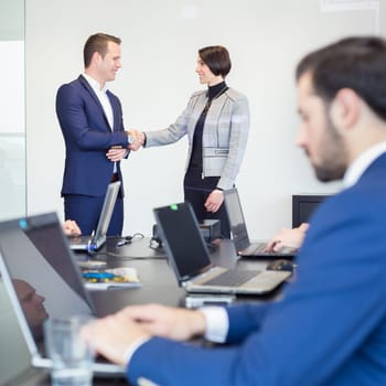 Sealing a deal. Business people shaking hands, finishing up meeting in corporate office. Businessman working on laptop in foreground. Business and entrepreneurship concept.