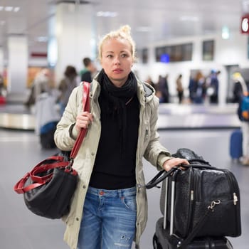 Casually dressed young female traveller walking in airport terminal hall with collected laggage after flight arrival . Blured background. Can also be used as railway, metro, bus station.