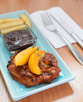 Grilled pork steak fillet with Peach and French fries on blue plate and cutlery over wooden table