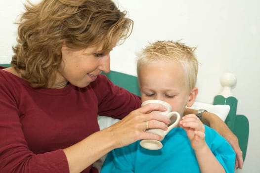 This boy drinks some coffee while lying relaxed in bed with his mommy.