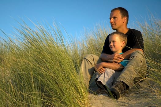 A boy and his father are looking far away, to the horizon. They are sitting in the dunes.
