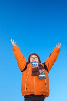Boy with wintercoat against a bright blue sky, throwing his hands up in the air.