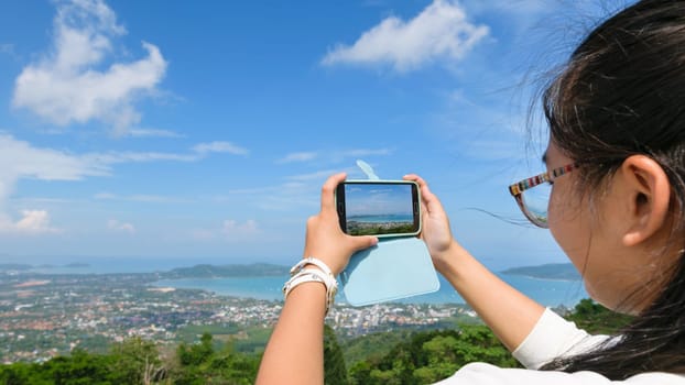 Young women tourist take photos high angle view of Ao Chalong bay and city sea side with mobile phones on top of Yot Khao Nak Kerd Hill in Phuket Province, Thailand, 16