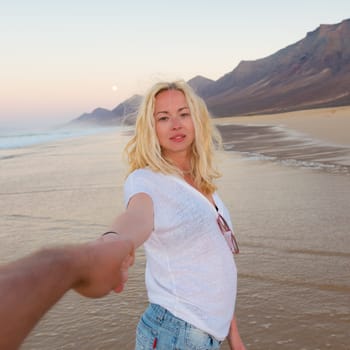 Young romantic couple, holding hands, having fun on perfect deserted beach at sunset. Shot from boyfrieds perspective. Guy looking at her beautiful carefree girlfriend.