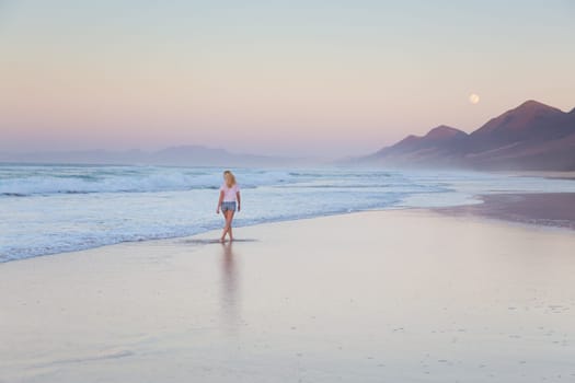 Woman walking on sandy beach in sunset. Waves sweeping away her traces in sand. Beach, travel, concept. Copy space.