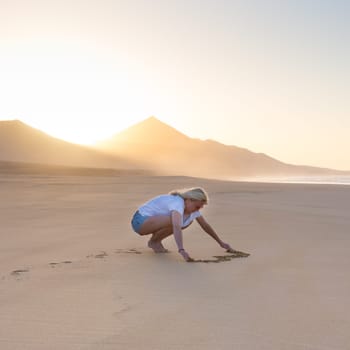Woman drawing heart in sand on beautiful solitary Cofete beach in sunset. Love, travel concept. Fuerteventura, Canary Islands, Spain.
