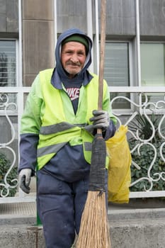 Tehran Province- IRAN-March 17, 2016 A Portrait of An Old Urban Street Cleaner Man Holding Hand-made Sweeping Tool By Gloved Hand in Front of Modern Building