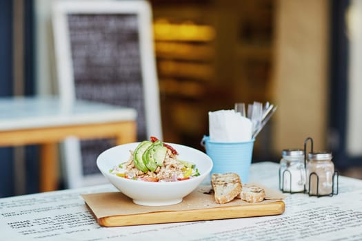 Fresh chcken and avacado sald in a presented in a bowl on a wooden board, alongside some freshly sliced bread laid out on a table outside a gourmet delicatessen eatery