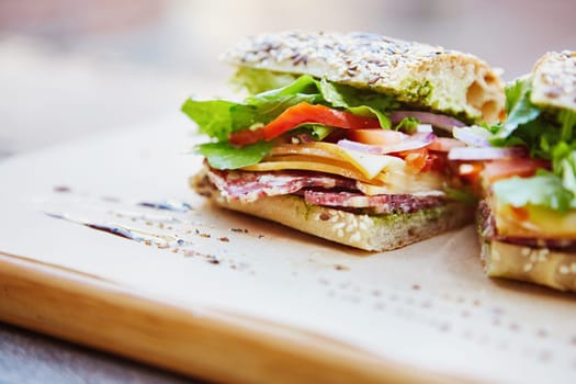 Healthy sandwich made of a fresh seeded roll, cut in half to display tasty ingredients of salami, tomato, lettuce and chees, presented on a wooden board