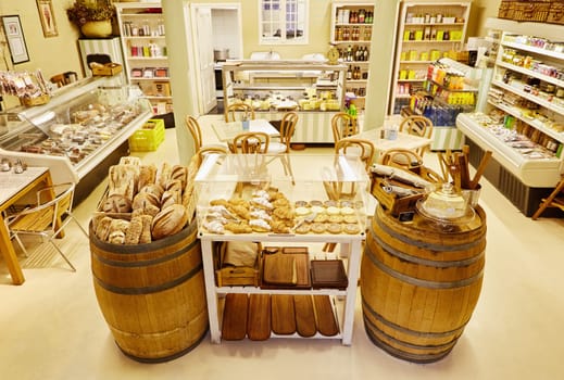 Interior view of a delicatessen with chairs and tables and displays featuring meats, preserves and general supplies as well a display of freshly baked breads and cakes in the foreground