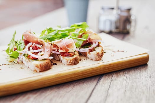 Gourmet meal of bruschetta topped with toamto, onion, parma ham and rocket, presented on a wooden board with condiments in the background