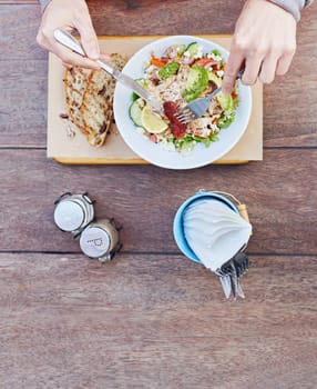 Overhead shot of a person&#39 s hands holding cutlery and about to start eating a fresh salad of chicken, avocado, sundried tomatoes and other ingredients on a wooden table with serviettes at the ready
