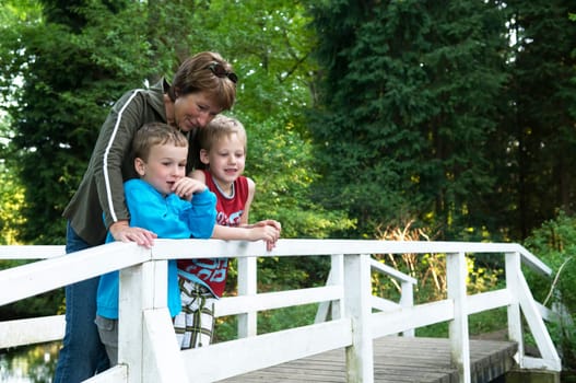 Grandma is standing on a bridge with her two grandsons, looking at a lake.