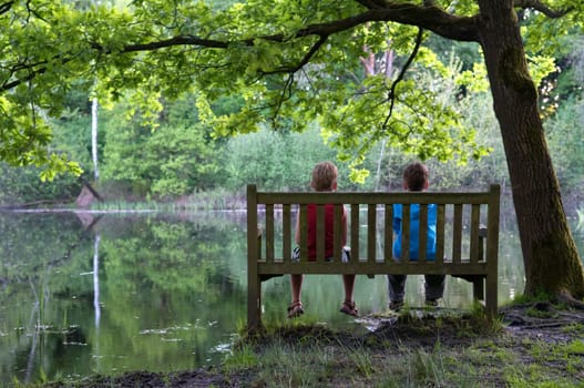 Two friends are sitting on a bench, looking at a lake.
