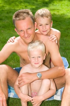 Happy family picture of a father and two sons sitting at the grass in summertime.