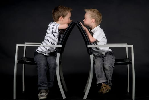 Two brothers sitting on a chair looking at each other, against a black background.