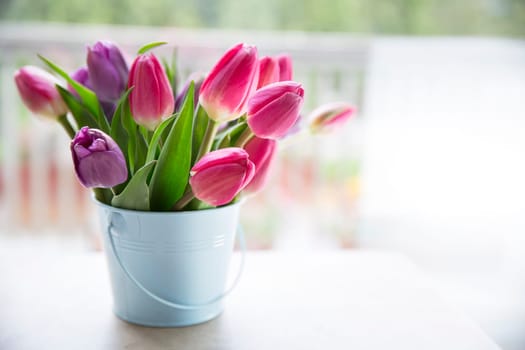 Bouquet of purple and pink tulips in light blue bucket in front of window.