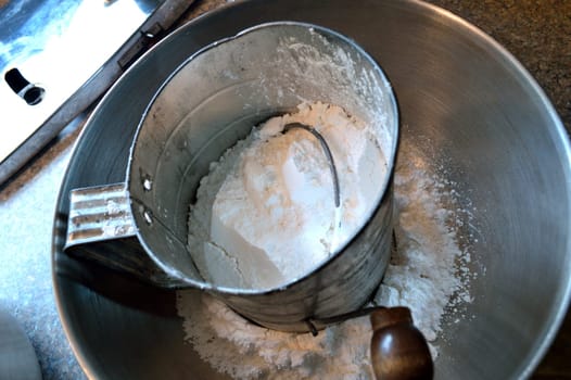 Overhead view of confectioner's (powdered) sugar in an old-fashioned hand sifter in a stainless steel bowl on countertop, with bottom of stand mixer to the left.