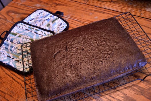 Dark chocolate cake sitting on a cooling rack to cool with colorful oven holders and sea green antique bowl with cake mix in it background on distressed wooden table in old-fashioned kitchen.