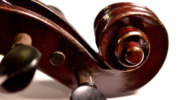 Letterbox sized image of an extreme closeup of an antique violin's scroll and tuning pegs against a white backdrop