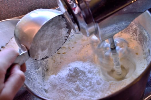 Adding 1 cup of powdered sugar to vanilla icing as its being mixed in a stainless steel stand mixer.