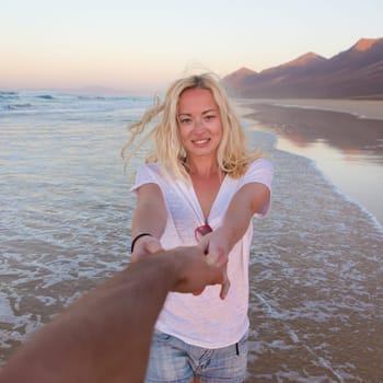 Young romantic couple, holding hands, having fun on perfect deserted beach at sunset. Shot from boyfrieds perspective. Guy looking at her beautiful carefree girlfriend.