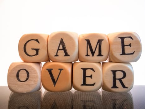 stacked wooden cubes with letters on glass plate result in the word GAME OVER in two rows on top of each other. White background.