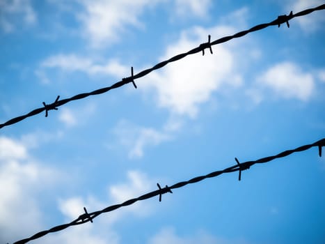 Two diagonally running barbed wires in front of blue sky with clouds.