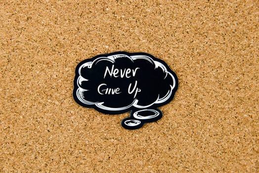 Never Give Up written on black thinking bubble over cork board background, copy space available