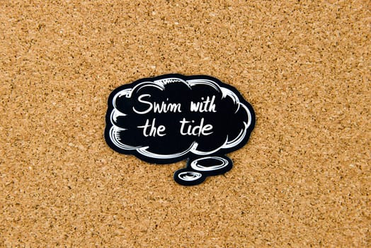 SWIM WITH THE TIDE written on black thinking bubble over cork board background, copy space available