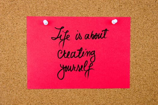 Red paper note with handwritten text Life Is About Creating Yourself pinned on cork board with white thumbtacks