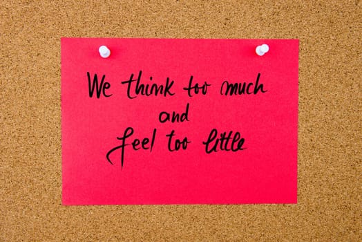 Red paper note with handwritten text We Think Too Much and Feel Too Little pinned on cork board with white thumbtacks
