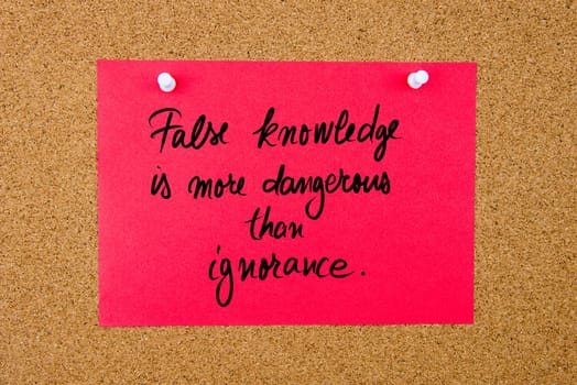 Red paper note with handwritten text False Knowledge Is More Dangerous Than Ignorance  pinned on cork board with white thumbtacks