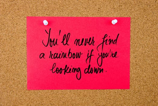 Red paper note with handwritten text You Will Never Find A Rainbow If You Are Looking Down pinned on cork board with white thumbtacks