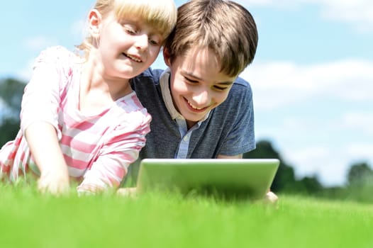 Brother and sister using tablet PC on the grass in summer park.