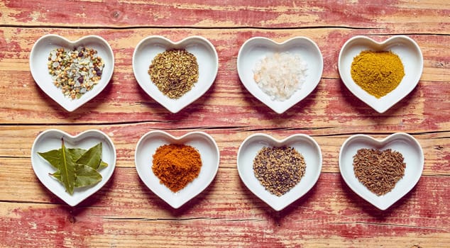 Assorted culinary spices and condiments in heart shaped dishes arranged in two rows on a rustic wood background, overhead view