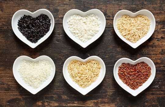 Selection of different rice grains in heart shaped dishes with par-boiled and husked white rice and long grain wild rice, overhead view on rustic wood