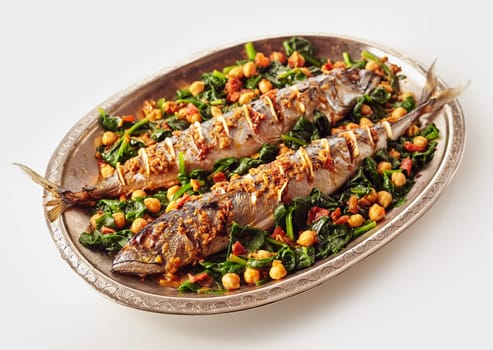 Single oval shaped silver platter full of two whole cooked mackerel fish stuffed with spices and garnished with beans and spinach