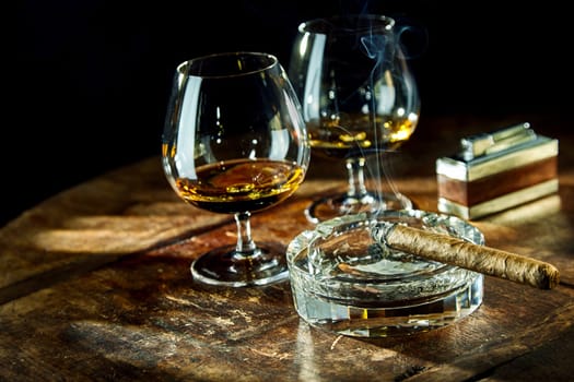 Two goblets of liquor besides smoking cigar sitting in glass ashtray by fancy lighter on a wooden table