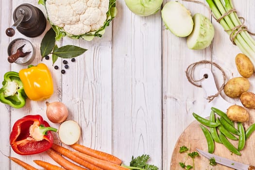 Delicious fresh vegetables forming a frame on white wood with central copy space together with a cutting board, knife and seasoning for cooking
