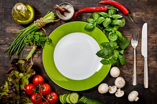 High Angle Still Life View of Green and White Plate with Knife and Fork on Rustic Wooden Table with Variety of Fresh Herbs and Healthy Raw Vegetables