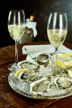 Pair of tall glasses of champagne next to silver platter of oysters and lemon slices on table with cooler and napkin in background