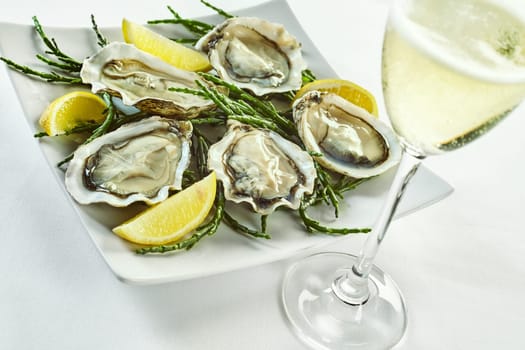Open oyster shells with lemon wedges in plate with curled edges and drink in tall champagne glass over white