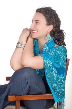 Young model in her mid twenties wearing aqua colored earrings of middle eastern design and a blue shawl sits on a wooden chair and smiles while looking in to the distance.