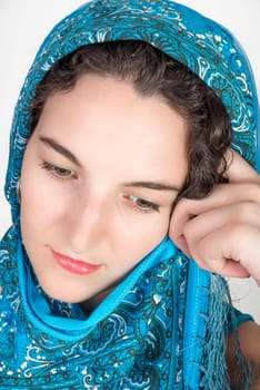 Young female model with black hair wearing a blue shawl over her hair held by left hand as shel looks down wards while thinking.