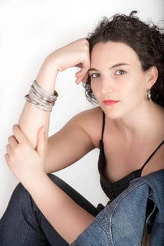 Young model in her mid twenties sitting on a chair with a blue denim jacket hanging over the back of the chair, head resting against hand, on white background.