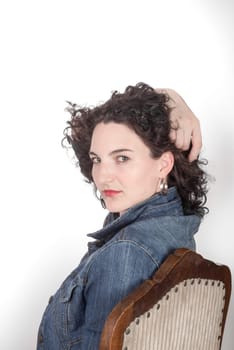 Portrait of a Young model in her mid twenties sitting on a wooden chair with a blue denim jacket, head turned to camera, and hand in hair, all in front of a white background.