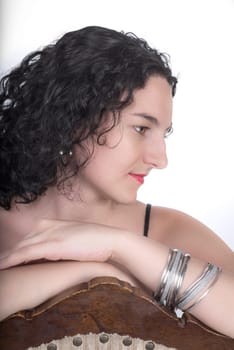 Young model in her mid twenties sitting on a chair with her arms on the back of the chair situated in front of a white background.