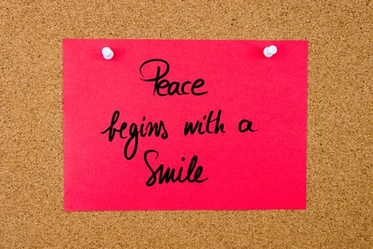 Red paper note with handwritten text Peace Begins With A Smile pinned on cork board with white thumbtacks